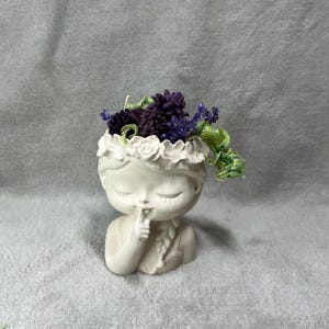 Peut inclure: Planteur en céramique blanche en forme de buste de fille avec une couronne de fleurs. Le planteur est rempli de fleurs séchées violettes et de feuillage vert.