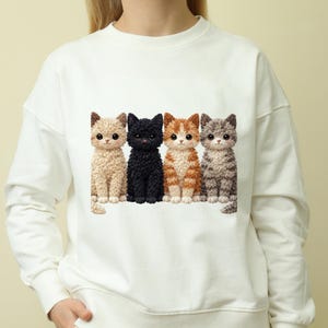 Könnte beinhalten: Weißes Sweatshirt mit einem Design von vier Cartoon-Katzen. Die Katzen sind beige, schwarz, orange und weiß sowie grau. Jede Katze hat große Augen und sitzt aufrecht. Das Sweatshirt ist ein lässiger Stil.