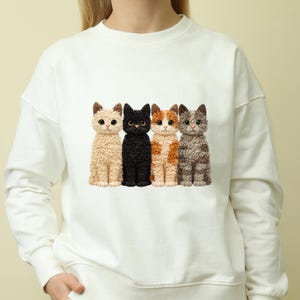 Könnte beinhalten: Weißes Sweatshirt mit einer Reihe von vier stilisierten Katzen. Die Katzen sind in verschiedenen Farben: Beige, Schwarz, Orange und Weiß sowie Grau. Die Katzen haben detaillierte Fellstrukturen und sitzen aufrecht. Das Sweatshirt hat einen Rundhalsausschnitt und lange Ärmel.