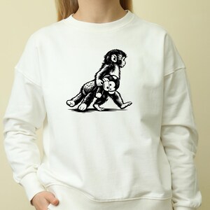 May include: White sweatshirt featuring a black and white illustration of a monkey carrying a stuffed animal. The monkey is walking, and the design is centered on the front of the sweatshirt. The sweatshirt has long sleeves and a crew neck.