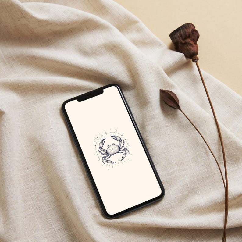 May include: A black smartphone displays a crab illustration within a circular design. The phone rests on a beige fabric, with dried plant elements nearby. The crab design is in a detailed, line-art style.