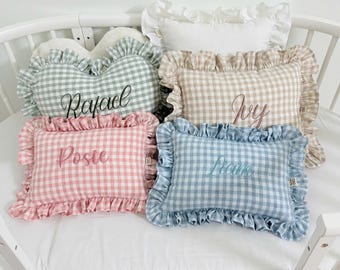 Può includere: Una collezione di cinque cuscini rettangolari con bordi a volant. Quattro cuscini presentano un motivo a quadretti vichy in verde salvia, beige, rosa e blu, ricamati con i nomi Rafael, Ivy, Posie e Liam. Un cuscino bianco tinta unita si trova dietro.