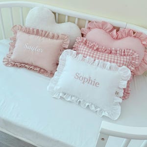 May include: A collection of rectangular and heart-shaped throw pillows with ruffled edges. One pink pillow features the embroidered name Saylor, while a white pillow features the embroidered name Sophie. Other pillows include pink gingham and polka dot patterns.