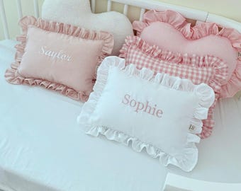 May include: A collection of decorative pillows in a crib. The pillows include a heart-shaped pillow, a pink gingham pillow, and two rectangular pillows with embroidered names. The pillows have ruffled edges and are in shades of pink and white.