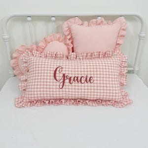 Peut inclure: Un coussin rectangulaire à motif vichy rose et blanc avec le prénom Gracie brodé en écriture cursive rose foncé. Le coussin présente un bord à volants et est accompagné de deux coussins roses plus petits avec des bords à volants.