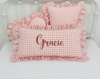 Può includere: Un cuscino rettangolare con motivo a quadretti vichy rosa e bianco con il nome Gracie ricamato in corsivo rosa scuro. Il cuscino presenta un bordo arricciato ed è accompagnato da due cuscini rosa più piccoli con bordi arricciati.