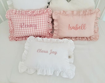 Può includere: Tre cuscini rettangolari con bordi arricciati. Uno è bianco con il nome Clara Jay ricamato in rosa. Uno è a quadretti vichy rosa con il nome Renny ricamato in rosa. Uno è rosa tinta unita con il nome Isabell ricamato in rosa.