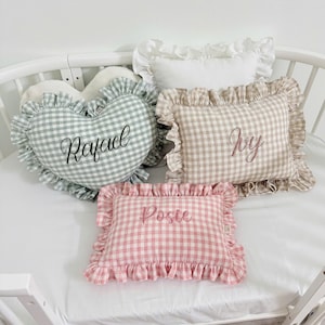 May include: A selection of personalised cushions on a cot. The cushions display gingham patterns in green, pink, and beige, with frilled edges. One cushion is heart-shaped, and others are rectangular. Each cushion has a name embroidered on it.