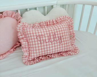 May include: A rectangular pillow with a pink and white gingham pattern, featuring a ruffled border and the name Tully embroidered in pink script in the center. A small white heart-shaped pillow is positioned behind the rectangular pillow.