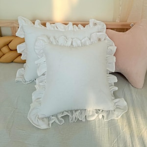 Könnte beinhalten: Zwei quadratische weiße Kissen mit gerüschten Rändern und einem kleinen Stoffetikett. Die Kissen sind auf einem Bett zusammen mit einem senfgelben geflochtenen Kissen und einem blassrosa sternförmigen Kissen angeordnet, was eine geschichtete Bettanordnung ergibt.