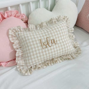 May include: A rectangular throw pillow featuring a beige and white gingham check pattern with a ruffled border. The name Isla is embroidered in a cursive brown script in the center of the pillow. A small fabric tag is attached to the side edge.