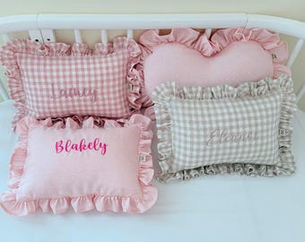 May include: Four rectangular and heart-shaped throw pillows with ruffled edges. One pink gingham pillow features the name Lainey, one solid pink pillow features the name Blakely, one beige gingham pillow features the name Eleanor, and one pink polka dot pillow.
