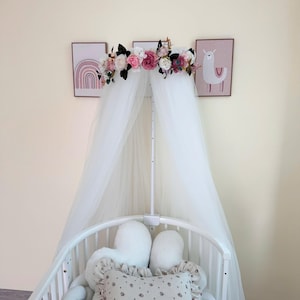 Può includere: Culla bianca con baldacchino, una ghirlanda di fiori, un baldacchino in tulle bianco e un cuscino bianco e beige con motivo floreale. La culla è decorata con due cuscini bianchi e soffici e una corda intrecciata bianca.