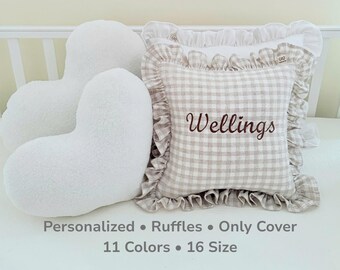 May include: A square throw pillow cover with a beige and white gingham pattern, featuring a ruffled border and the embroidered name "Wellings" in brown script. Beside it are two white heart-shaped plush pillows with a textured, fuzzy surface.