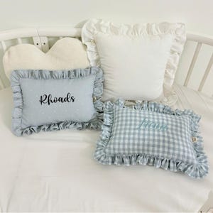 May include: Four decorative pillows in a crib. One white pillow with ruffled edges, one light blue pillow with white polka dots and the name "Rhoads", and one blue and white checkered pillow with the name "Lucie". A heart-shaped pillow is also visible.