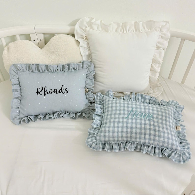 May include: Four decorative pillows in a crib. One white pillow with ruffled edges, one light blue pillow with white polka dots and the name "Rhoads", and one blue and white checkered pillow with the name "Lucie". A heart-shaped pillow is also visible.