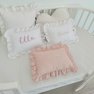 May include: White and pink ruffled pillows with embroidered names. The pillows are rectangular and square, with the names "Ella", "Sloane", and "Jelaiynn" embroidered in cursive. A cloud-shaped pillow is also present.