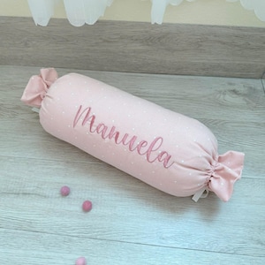 May include: A cylindrical bolster pillow in light pink fabric with a white polka dot pattern. The name Manuela is embroidered in cursive script on the center. The ends of the pillow are gathered and tied with fabric, resembling a candy wrapper.