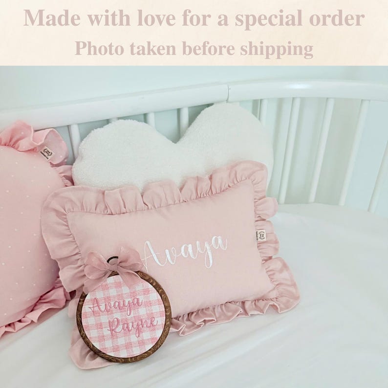 May include: Pink decorative pillows with ruffled edges and embroidered names. One pillow is rectangular with the name "Avaya" embroidered in white. A round embroidery hoop with the name "Avaya Rayne" is also visible.
