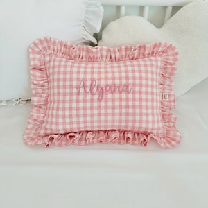May include: A rectangular throw cushion featuring a pink and white gingham check pattern with a ruffled border. The centre of the cushion is embroidered with the name Alyana in a cursive script font. A small fabric tag is attached to the right side.