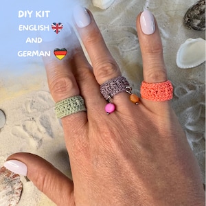 Könnte beinhalten: Eine Hand zeigt drei gehäkelte Ringe in Grün, Lila und Orange. Der lila Ring hat einen rosa Perlenanhänger. Das Bild enthält den Text "DIY KIT ENGLISH AND GERMAN".