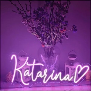Pode incluir: Uma placa de néon com o nome "Katarina" em fonte cursiva e um símbolo de coração, brilhando com uma luz rosa suave. A placa está sobre um fundo roxo com elementos decorativos.