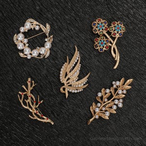 Puede incluir: Cinco broches dorados con varios diseños se muestran sobre una superficie oscura. Los broches incluyen una corona de perlas, un diseño floral con gemas de colores, una hoja con perlas y una rama con detalles rojos.