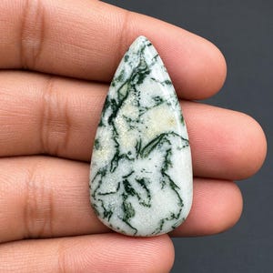 Può includere: Una gemma cabochon a forma di pera caratterizzata da una base bianca con intricate inclusioni verde scuro simili a muschio su tutta la superficie. La pietra presenta una finitura liscia e lucida con una distinta silhouette a goccia.