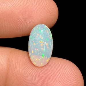 May include: An oval-shaped cabochon gemstone with a translucent white base and a play-of-color featuring scattered flecks of bright green, orange, and yellow light. The stone has a smooth, polished surface and is held against a black background.
