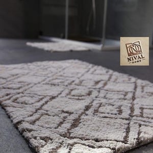 Könnte beinhalten: Ein rechteckiger, hellfarbiger Badteppich mit geometrischem Muster. Der Teppich besteht aus einem weichen, plüschigen Material und weist ein sich wiederholendes quadratisches und rechteckiges Design in Beige- und Brauntönen auf. Das Logo "NIVAL HOME" ist sichtbar.