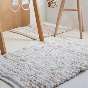 Op de afbeelding: Een rechthoekige badmat in wit en bruin, met een textuur van bolletjes. De mat ligt op een lichtgekleurde tegelvloer. Een houten handdoekenrek en krukje zijn zichtbaar op de achtergrond.