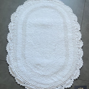 Peut inclure: Un tapis de bain ovale blanc doté d'une zone centrale texturée et d'une bordure décorative en crochet avec un bord festonné. Le tapis présente une couleur blanche uniforme et inclut un petit logo dans le coin portant l'inscription NIVAL HOME.