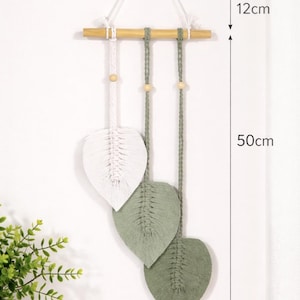 Può includere: Applique da parete in macramè con tre disegni a forma di foglia in bianco e verde salvia, sospeso a un tassello di legno. Il pezzo misura 20 cm di larghezza, 50 cm di lunghezza e 12 cm di altezza. Arredamento per la casa.