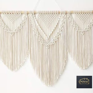 Könnte beinhalten: Cremefarbener Makramee-Wandbehang mit einem Holzstab. Das Design besteht aus drei Abschnitten mit aufwendigen Knoten und Fransen. Der Mittelteil hat ein V-förmiges Design, und die beiden anderen Abschnitte haben ein ähnliches, aber kleineres Design. Ein dekoratives Wohnaccessoire.