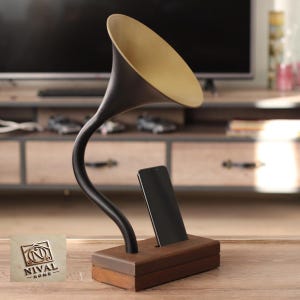 Puede incluir: Un soporte decorativo para teléfono con forma de gramófono antiguo. Cuenta con una base de madera marrón oscuro, un brazo negro curvado y una bocina dorada. Un smartphone negro está colocado en el soporte. El logotipo "NIVAL - HOME" es visible en una placa de madera.