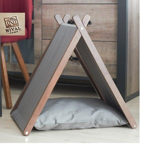May include: A wooden pet house in a teepee shape with a gray cushion inside. The frame is made of brown wood with gray panels. The cushion is a light gray color. The NIVAL HOME logo is visible in the upper left corner.