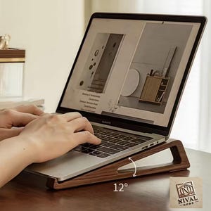 Op de afbeelding: Een donkerbruine houten laptopstandaard, onder een hoek van 12 graden, ter ondersteuning van een laptop. De standaard heeft een uitsparing en het laptopscherm toont een webpagina. De standaard is voorzien van de merknaam "NIVAL HOME".