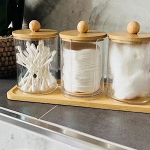 Op de afbeelding: Drie glazen potten met bamboe deksels en een houten dienblad. De potten bevatten wattenstaafjes, wattenschijfjes en wattenbolletjes. Op een grijze aanrecht met een plant op de achtergrond.
