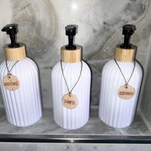 Könnte beinhalten: Drei weiße, geriffelte Seifenspender mit Bambusdeckeln und schwarzen Pumpen. Jede Flasche hat ein Holzschild an einer Kette mit der Aufschrift "Conditioner", "Shampoo" und "Bodywash". Die Spender sind auf einem Marmorregal angeordnet.