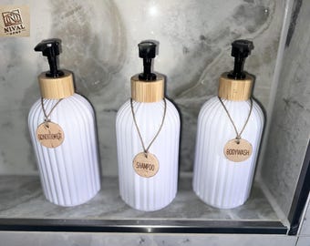 May include: Three white, ribbed, refillable soap dispensers with bamboo lids and black pumps. Each bottle has a wooden tag on a chain, labeled "Conditioner", "Shampoo", and "Bodywash". The dispensers are arranged on a marble shelf.