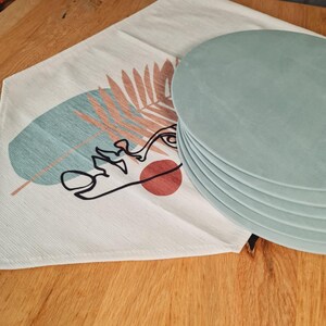 Könnte beinhalten: Ein Stapel von sechs hellblauen runden Tischsets neben einem weißen Tischläufer. Der Tischläufer hat ein Design aus einem Gesicht, einem Blatt und einem roten Kreis. Das Design ist in Schwarz, Türkis und Pfirsich gehalten.