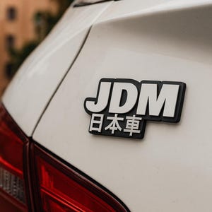 May include: A black and white JDM (Japanese Domestic Market) emblem is affixed to the white rear of a car. The emblem features the letters "JDM" above Japanese characters. The car's red taillight is visible.