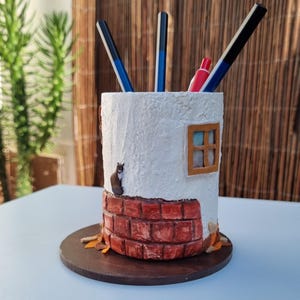 May include: A handmade white pen holder with a textured finish, featuring a brick base and a small window. A cat figurine sits on the side. The holder contains pens and a red marker. It rests on a round, dark brown wooden base.