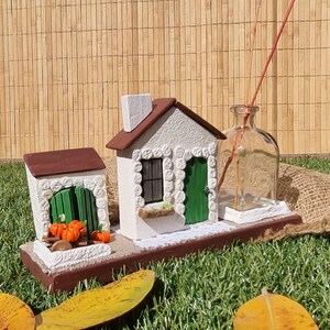 Może przedstawiać: Dekoracyjna miniaturowa scena wioski z dwoma białymi budynkami z brązowymi dachami i zielonymi drzwiami. Małe pomarańczowe dynie umieszczono przed jednym z budynków. W zestawie znajduje się również przezroczysta szklana butelka z trzciną. Scena jest osadzona na brązowej podstawie.