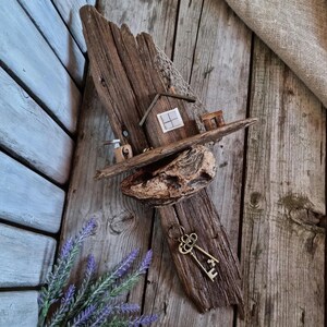 Peut inclure: Une œuvre d'art murale rustique en bois représentant une maison miniature avec une petite fenêtre, un banc et une clé. L'œuvre est faite de bois flotté et d'autres matériaux naturels, avec un aspect vieilli. Les clés sont en laiton.