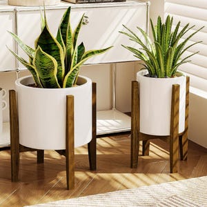 May include: Two white ceramic plant pots with green plants, set on wooden stands. The pots have a cylindrical shape and are elevated by four brown wooden legs. The plants have green and yellow leaves, adding a natural touch to the decor.