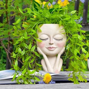 May include: A cream-coloured face planter with closed eyes, rosy cheeks, and long eyelashes, supporting its head with its hands. The planter is filled with vibrant green foliage and yellow and blue flowers, creating a whimsical garden accent.