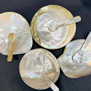 May include: Four iridescent mother-of-pearl dishes with spoons. Two dishes have gold-coloured fish designs on the rims. The spoons and dishes have a pearlescent sheen, reflecting light. The dishes are arranged on a dark surface.