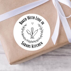May include: A round sticker on a wrapped parcel with the text "Baked With Love In Sarahs Kitchen". The sticker features a whisk, spatula, and rolling pin inside a wreath. The parcel is wrapped in brown paper and tied with a white ribbon.
