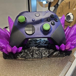 May include: A purple and black video game controller sits in a 3D printed stand with purple crystal-like accents. The stand has the text "Driftpete" printed on it.